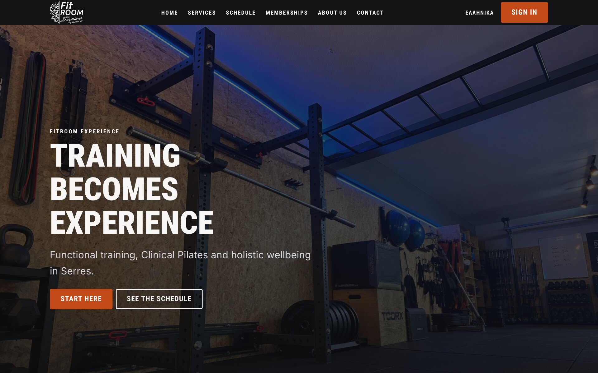 FitRoom Experiences forside: en mørk topbjælke med det hvide FitRoom-logo, linkene Home, Services, Schedule, Memberships, About us og Contact, et sproglink "Ελληνικά" og en orange "Sign in"-knap; nedenunder et mørkt foto af studiet, et sort rack med en vægtstang under en klatrestige i loftet foran blåt oplyste spånpladevægge, med overskriften "Training becomes experience", linjen "Functional training, Clinical Pilates and holistic wellbeing in Serres." og to knapper, en orange "Start here" og en "See the schedule" med kant.