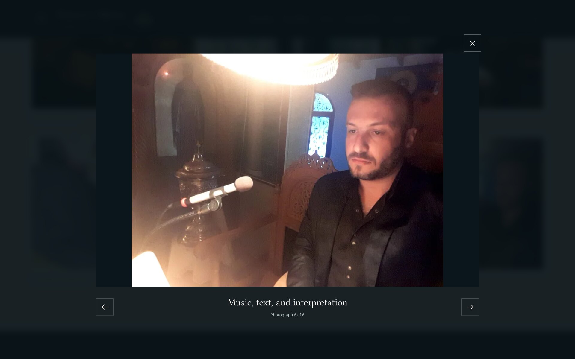 The photo viewer open over the dimmed gallery: a photograph of Petros Nikitas seated beside a microphone under a bright lamp, a close button at the top right, previous and next arrows below, and between them the caption "Music, text, and interpretation" with "Photograph 6 of 6".