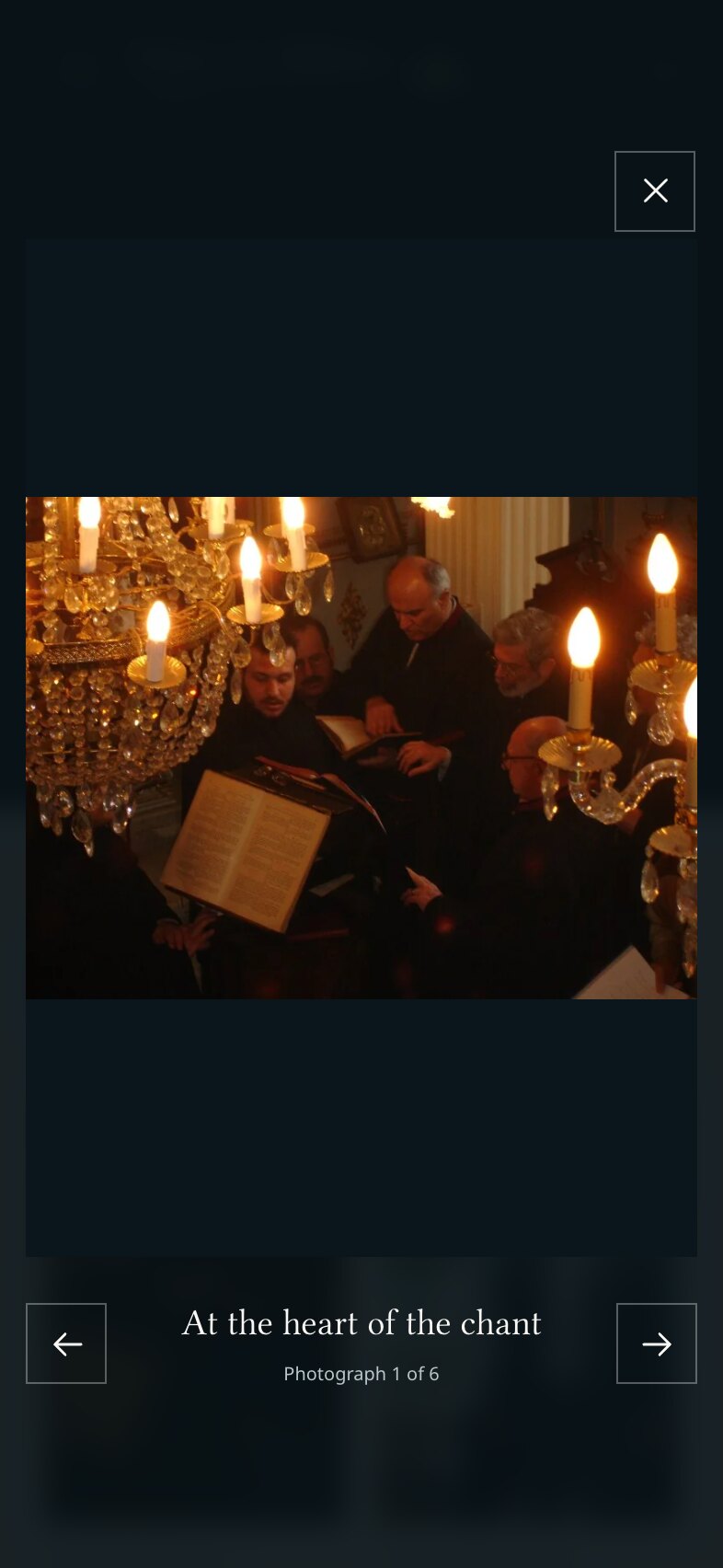 The photo viewer on a phone: a photograph of chanters reading a score by candlelight beneath a chandelier, a close button above it, previous and next arrows below and between them the caption "At the heart of the chant" with "Photograph 1 of 6".