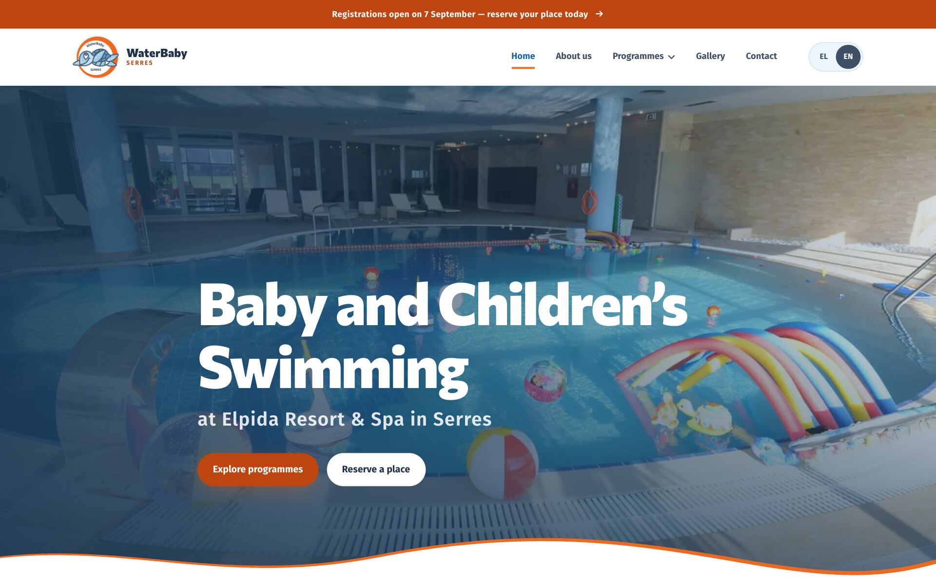 WaterBaby 首页:橙色公告栏和导航栏下方,语言切换选中 EN;标题“Baby and Children’s Swimming”和一行“at Elpida Resort & Spa in Serres”叠在一张调暗的室内泳池照片上,池中有彩虹色充气拱门、沙滩球和漂浮玩具;下方是橙色按钮“Explore programmes”、白色按钮“Reserve a place”,以及一条橙色描边的波浪。